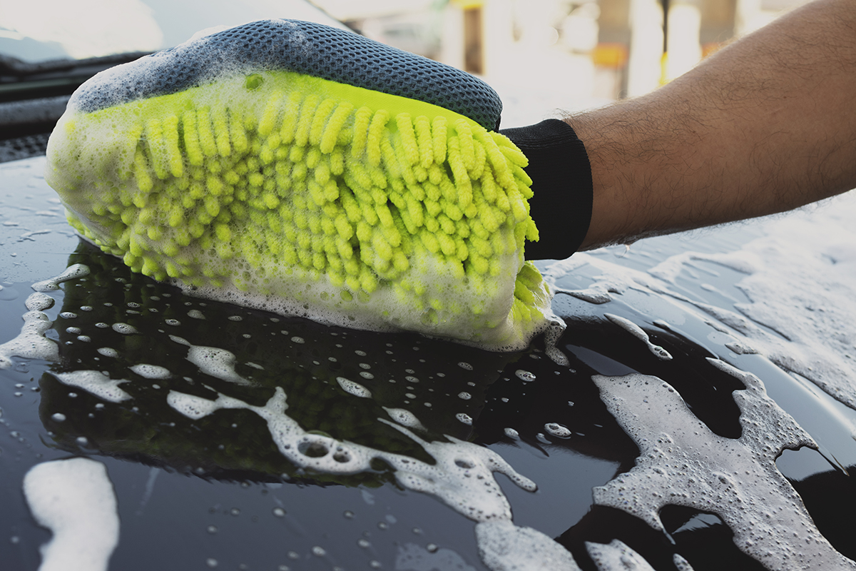Mano de un hombre con un guante de microfibra amarillo limpiando el capó de un coche lleno de espuma