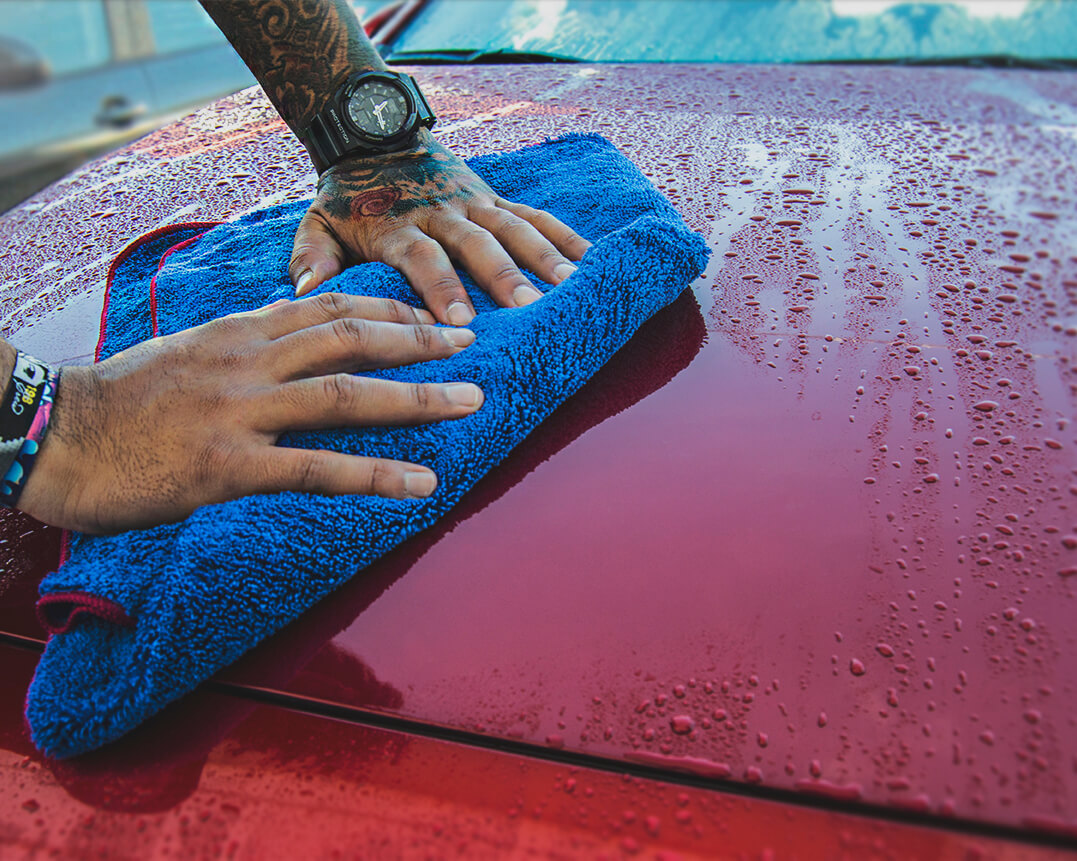 Manos de hombre con tatuajes secando el capó de un coche rojo con una toalla de secado azul
