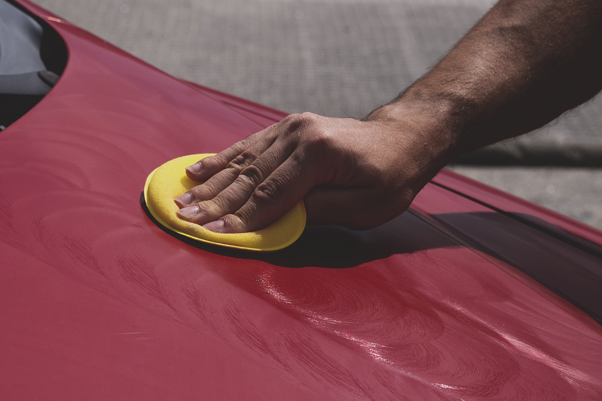 Mano de un hombre con un aplicador de poliespuma amarillo sobre el capó de un coche rojo