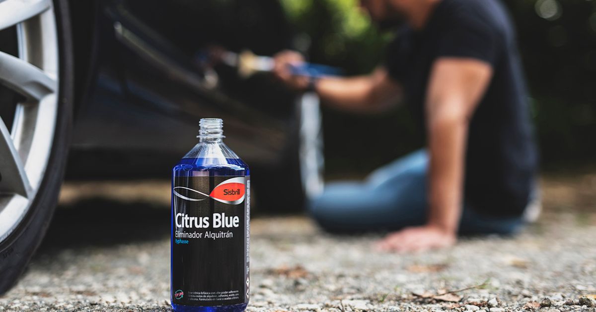  Una botella de eliminador de alquitrán Sisbrill Citrus Blue está en primer plano en el suelo junto a la rueda de un coche, mientras que una persona en el fondo, ligeramente desenfocada, parece estar aplicando el producto al vehículo con un pincel.