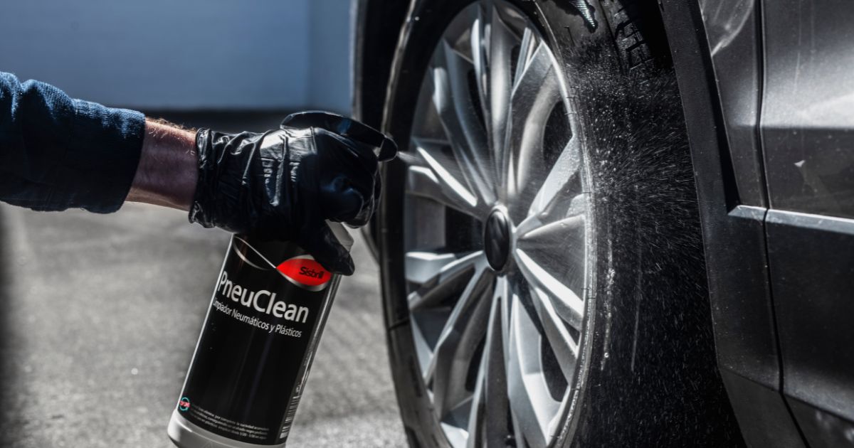 Una persona con guantes negros está rociando un neumático con un producto llamado "PneuClean" de la marca Sisbrill, diseñado para limpiar neumáticos y plásticos. El spray crea una neblina al ser aplicado sobre la rueda del coche, indicando un proceso de limpieza activo.