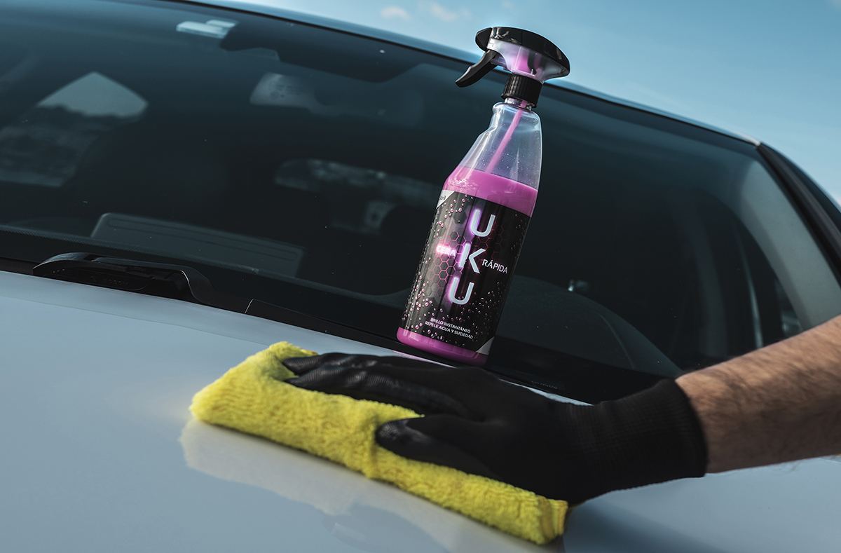 Botella de UKU Cera Rápida encima del capó de un coche blanco mientras una mano con un guante negro de un hombre desliza una microfibra amarilla