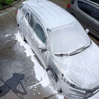 Coche plateado completamente cubierto de espuma durante un lavado, visto desde arriba, con una persona usando una manguera a presión visible en la sombra sobre el pavimento.