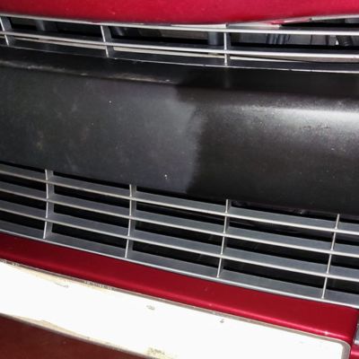 Parrilla frontal de un coche de color rojo con una parte plástica negra que muestra una sección más limpia comparada con el resto.